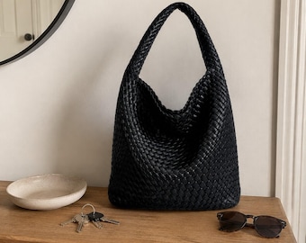 May include: A black hobo bag featuring a woven texture and a single integrated shoulder strap. The bag has a slouchy, unstructured silhouette and is positioned on a wooden surface alongside a small bowl, a set of keys, and sunglasses.