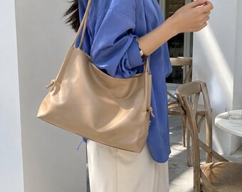 May include: A beige shoulder bag with a smooth, matte finish and a rectangular silhouette. The bag features thin, matching shoulder straps and side detailing with small knotted accents at the top corners, creating a soft, unstructured shape.