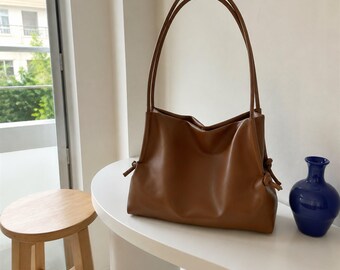 May include: A medium-sized, smooth, brown shoulder bag with a soft, slouchy structure. The bag features two long, thin, matching brown shoulder straps and decorative knotted cord details on both sides of the main compartment opening.