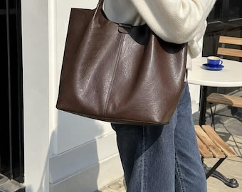 May include: A dark brown tote bag with a smooth, slightly textured finish. The bag features a vertical seam down the center, a wide opening, and two shoulder straps. The material has a subtle sheen, and the design is simple and functional.