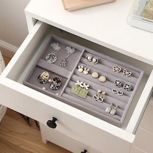 May include: A drawer filled with a gray jewelry organizer holding various earrings. The earrings include pearl studs, bow-shaped earrings, and floral designs. The drawer is part of a white cabinet.