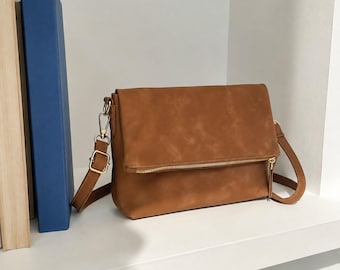 May include: A tan fold-over crossbody bag with a smooth matte finish, featuring a gold-tone metal zipper along the front flap, matching tan shoulder strap with an adjustable gold-tone buckle, and gold-tone lobster claw clasps on both sides.