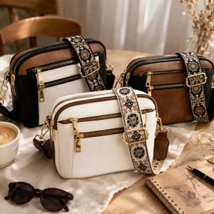May include: Three crossbody bags in black, brown, and white with gold zippers and decorative straps. The bags have a rectangular shape with a top zipper closure. The straps feature a floral pattern. Sunglasses and a cup of coffee are in the foreground.