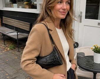 May include: A camel-colored wool coat worn over a white ribbed top, paired with a black crocodile-embossed leather shoulder bag and dark trousers. The bag features a curved silhouette and a thin strap, while the coat has a classic lapel collar.