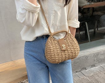 May include: A woven straw handbag with a rounded shape, featuring a tan leather-like center strap with a gold-tone metal turn-lock closure. The bag includes a matching woven straw top handle and a thin, long woven straw shoulder strap.