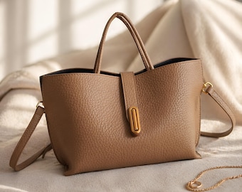 May include: A tan leather handbag with a textured surface. The bag has two handles and a detachable shoulder strap. A gold-colored clasp secures the front flap. The bag is sitting on a cream-colored surface.