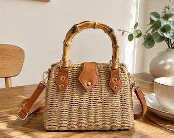 May include: A structured handbag crafted from woven tan straw, featuring a curved bamboo handle and a tan faux leather flap closure with a gold-tone snap button. The bag includes tan faux leather side attachments with gold rivets and a matching shoulder strap.