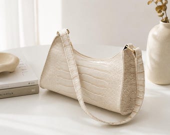 May include: A cream-colored shoulder bag with a textured crocodile-embossed pattern. The bag features a curved top silhouette, a single fixed shoulder strap, and gold-toned metal hardware attachments connecting the strap to the main body.