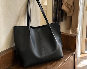 May include: A black tote bag with a smooth, matte finish, featuring two long shoulder straps and a vertical seam running down the center of the front panel. The bag has a rectangular shape with a wide opening and a structured base.