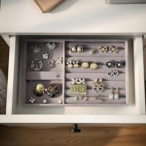 Puede incluir: Un cajón abierto revela un organizador de joyas gris lleno de varios pendientes, collares y broches. Las joyas incluyen piezas de perlas, oro y plata, con diseños florales y geométricos. El cajón forma parte de una cómoda blanca.