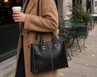 May include: A structured black leather handbag with dual top handles and a detachable shoulder strap. The bag features visible white contrast stitching, silver-toned metal hardware, and a rectangular silhouette. It is worn with a camel-colored coat.