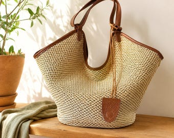 May include: A beige woven straw tote bag featuring brown leather trim along the top edges and handles. A thin leather cord hangs from one handle, attached to a small, textured brown leather tag. The bag has a wide, open-top design.