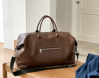 May include: A dark brown duffel bag featuring a smooth finish, two top handles, and a front exterior pocket with a silver-toned zipper closure. The bag includes a detachable black shoulder strap with silver-toned metal hardware clips on each side.