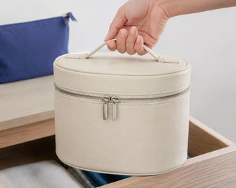 May include: An oval-shaped cream-colored vanity case with a top handle and a double-zipper closure. The case features a smooth, solid-colored exterior and a structured design, providing a compact storage solution for various small personal items.