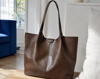 May include: A dark brown tote bag with a textured finish, featuring two long shoulder straps, a central vertical seam, and a small tab closure at the top center. The bag has a wide, open-top design with a structured yet flexible appearance.