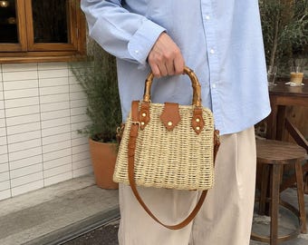 May include: A structured straw handbag featuring a woven beige body, two curved bamboo handles, and brown leather accents including a central tab closure and an adjustable shoulder strap with gold-toned metal hardware and rivets.
