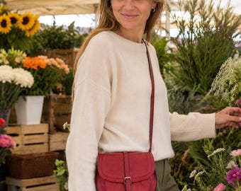May include: A red leather crossbody bag with a rounded flap, a central vertical strap, and a gold-toned metal buckle closure. The bag features a long, thin matching red shoulder strap and is worn over a cream-colored long-sleeved knit sweater.