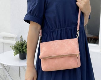 May include: A light pink fold-over crossbody bag with a gold-toned metal zipper closure along the front flap. The bag features a matching pink adjustable shoulder strap attached with gold-toned metal clips on each side of the rectangular body.