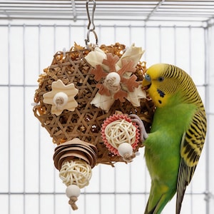 Puede incluir: Un periquito verde y amarillo junto a un juguete colgante para pájaros. El juguete está hecho de materiales naturales tejidos, con detalles de madera y papel. El pájaro sostiene una pequeña bola con su pata.
