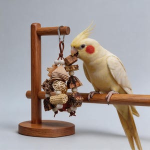 Puede incluir: Un cacatúa amarilla con una mancha roja en la mejilla se posa en un soporte de madera, interactuando con un juguete colgante para pájaros. El juguete presenta varios elementos de madera y tejidos, diseñados para el enriquecimiento aviar.