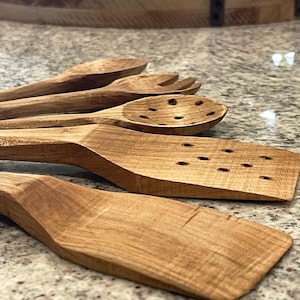 May include: A collection of wooden kitchen utensils, including a slotted spoon, spatula, and serving fork. The utensils are made of light brown wood and are arranged on a speckled surface. The wooden spoons and spatula have a natural wood grain.