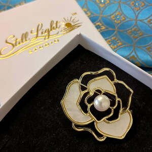 May include: A gold-toned metal and mother-of-pearl flower brooch with a pearl center, presented in a white box. The box has gold lettering that reads "Still Light Studio". The brooch is on a black surface.
