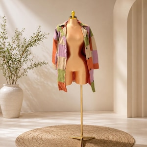 May include: A long-sleeved button-down shirt featuring a patchwork design with square panels in shades of lavender, olive green, and burnt orange. The shirt is displayed on a metallic gold-toned mannequin torso with a central stand.