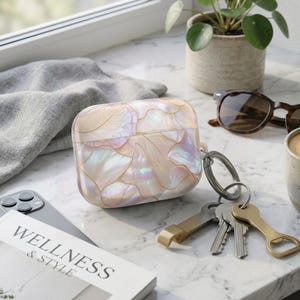 Puede incluir: Una funda protectora rectangular para auriculares inalámbricos con un patrón de concha iridiscente y nacarado con contornos dorados. La funda incluye una anilla metálica plateada con un juego de llaves y un abridor de botellas dorado.