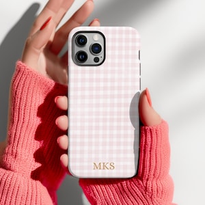Puede incluir: Una funda para smartphone con un patrón de cuadros vichy en rosa claro y blanco. La funda tiene un recorte para un sistema de triple cámara e incluye las letras de monograma MKS en color dorado impresas cerca del centro inferior.