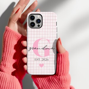 Puede incluir: Una funda para smartphone con un patrón de cuadros vichy en rosa claro y blanco. El diseño incluye una letra G grande en rosa, la palabra grandma en letra cursiva negra, el texto EST. 2026 y un pequeño icono de corazón rosa centrado.