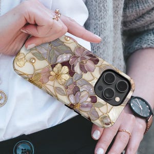 Op de afbeelding: Een smartphonehoesje met een bloemenpatroon ge&iuml;nspireerd op glas-in-lood in cr&egrave;me, mauve en goudtinten. Het ontwerp bevat ingewikkelde bloemblaadjes met gouden omlijningen en een uitsparing voor de drievoudige camera rechtsboven.
