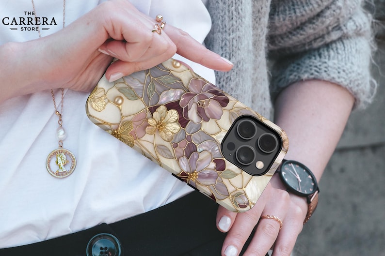 Op de afbeelding: Een smartphonehoesje met een bloemenpatroon ge&iuml;nspireerd op glas-in-lood in cr&egrave;me, mauve en goudtinten. Het ontwerp bevat ingewikkelde bloemblaadjes met gouden omlijningen en een uitsparing voor de drievoudige camera rechtsboven.