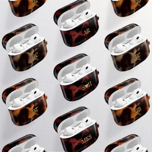 Może przedstawiać: Kolekcja otwartych, spersonalizowanych etui na słuchawki AirPod. Etui mają wzór w kolorze szylkretu w odcieniach brązu i czerwieni. Każde etui zawiera białe słuchawki i złote inicjały. Etui są ułożone na białym tle.