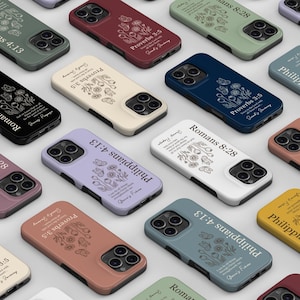 Puede incluir: Una colección de fundas para smartphone en varios colores sólidos, incluyendo negro, blanco, verde salvia, azul marino, lavanda, amarillo mostaza y terracota. Cada funda presenta una ilustración floral de líneas y un versículo bíblico.