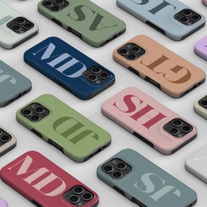 Puede incluir: Una colección de fundas rectangulares para teléfonos inteligentes dispuestas en una cuadrícula. Cada funda presenta un color sólido único y un monograma grande y llamativo de dos letras impreso en un tono contrastante.