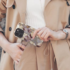Op de afbeelding: Een smartphonehoesje met een bloemenpatroon in glas-in-loodstijl met paarse en witte bloemblaadjes, goudkleurige omlijningen en een zwarte uitsparing voor de cameralens. Het hoesje heeft een glanzende afwerking en wordt vastgehouden tegen een beige trenchcoat. Het Carrera Store logo is zichtbaar.
