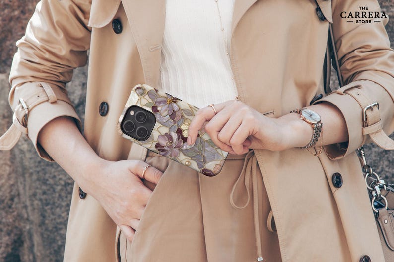 Op de afbeelding: Een smartphonehoesje met een bloemenpatroon in glas-in-loodstijl met paarse en witte bloemblaadjes, goudkleurige omlijningen en een zwarte uitsparing voor de cameralens. Het hoesje heeft een glanzende afwerking en wordt vastgehouden tegen een beige trenchcoat. Het Carrera Store logo is zichtbaar.