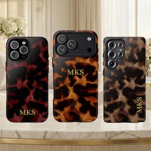 Puede incluir: Tres fundas para teléfono con un diseño de carey en tonos rojos, marrones y beige. Cada funda tiene una configuración de cámara diferente y las letras doradas "MKS". Las fundas se exhiben sobre una superficie dorada.