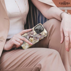 Op de afbeelding: Een smartphonehoesje met een bloemenpatroon in glas-in-loodstijl in cr&egrave;me, goud en paarse tinten. Het ontwerp bevat gedetailleerde omlijningen en kleine metalen studs. De tekst THE CARRERA STORE is zichtbaar in de rechterbovenhoek.