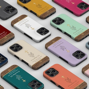 Puede incluir: Una colección de fundas para smartphone en varios colores sólidos, cada una con un acento de veta de madera en la parte superior e inferior. Las fundas tienen un gráfico floral, una cruz dorada y el texto Proverbs 3:5 Trust in the Lord with all thine heart.