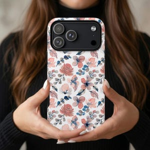 Puede incluir: Una funda de teléfono blanca con un estampado floral en tonos rosa, melocotón y azul. La funda presenta un diseño de acuarela repetido de flores y hojas. La funda del teléfono se sostiene frente a una persona.