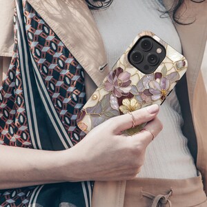 Op de afbeelding: Een smartphonehoesje met een bloemenpatroon in glas-in-loodstijl in paarse, roze en gouden tinten op een cr&egrave;mekleurige achtergrond. Het ontwerp bevat gedetailleerde donkere omlijningen en een uitsparing voor de camera in de linkerbovenhoek.