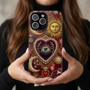 Puede incluir: Una funda para smartphone con fondo rojo intenso, un corazón morado central con borde de perlas y un motivo de ojo verde. El diseño incluye soles dorados, lunas crecientes, estrellas, una granada y un corazón anatómico rojo.