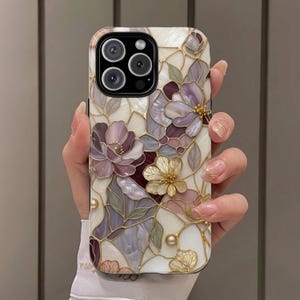 Op de afbeelding: Een smartphonehoesje met een ontwerp in glas-in-loodstijl, voorzien van iriserende bloempatronen in tinten paars, cr&egrave;me en goud. Het oppervlak toont een moza&iuml;ek van bloemblaadjes en bladeren met gouden omlijningen, geaccentueerd met kleine parelmoerachtige kralen.
