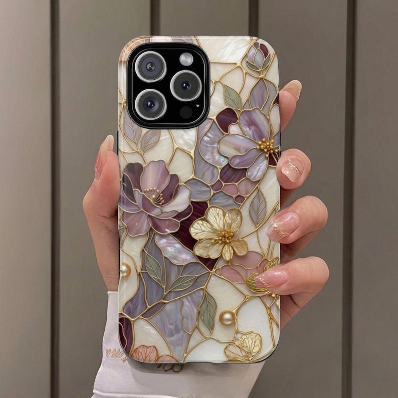Op de afbeelding: Een smartphonehoesje met een ontwerp in glas-in-loodstijl, voorzien van iriserende bloempatronen in tinten paars, cr&egrave;me en goud. Het oppervlak toont een moza&iuml;ek van bloemblaadjes en bladeren met gouden omlijningen, geaccentueerd met kleine parelmoerachtige kralen.
