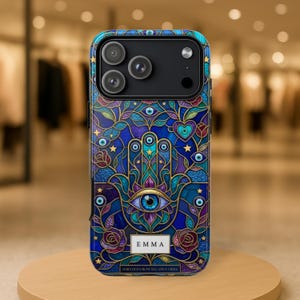May include: A blue and gold phone case featuring a Hamsa hand design with an eye in the center. The case has a stained glass effect with floral and star accents. The name "EMMA" is displayed on the case, along with the text "SHIELDED FROM NEGATIVE VIBES."