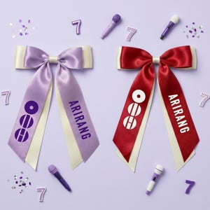 May include: Two satin ribbon bows, one lavender and one red, with cream-colored accents. Each bow has long streamers with the word "ARIRANG" printed on them. The lavender bow has a circular graphic, and the red bow has a different circular graphic. Miniature microphones and the number 7 are scattered around.