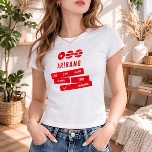Könnte beinhalten: Weißes T-Shirt mit rotem Text und Grafiken. Das Wort "ARIRANG" ist prominent dargestellt, zusammen mit anderen Namen. Das Shirt hat einen Rundhalsausschnitt und kurze Ärmel.