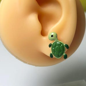 May include: A green turtle stud earring is shown on a mannequin ear. The turtle has a dark green shell and head, with black detailing. The earring is set against a light yellow background.