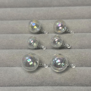 May include: Six clear, iridescent glass earrings with a bubble-like design. Each earring features a small, round glass bead connected to a post. The earrings are displayed on a gray, textured surface.